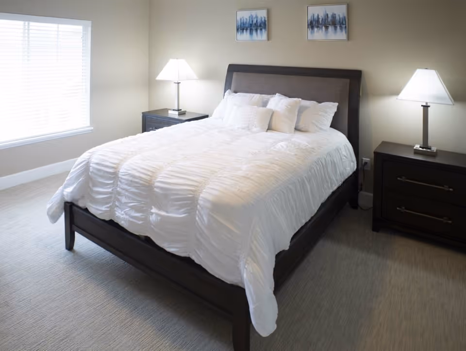 A neatly made bed with a white comforter and multiple pillows in a bedroom. The bed has a dark wooden frame and headboard. There are two matching dark wooden nightstands on either side of the bed, each with a modern table lamp. Two framed abstract cityscape paintings hang on the wall above the headboard. A large window with white blinds allows natural light into the room.