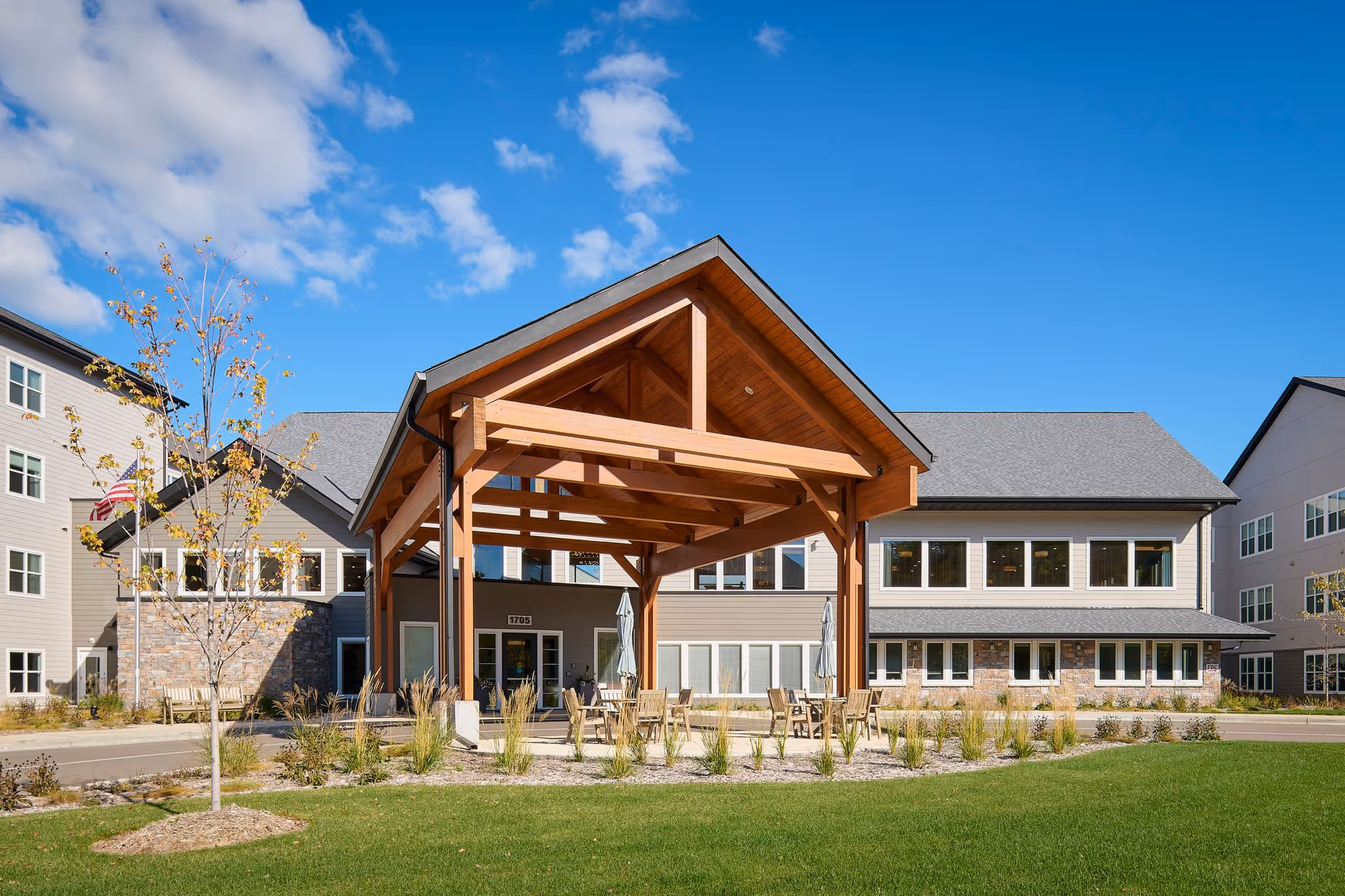 Front entrance of a modern senior living building with a wooden porte-cochère, patio seating, and landscaped lawn under a blue sky.
