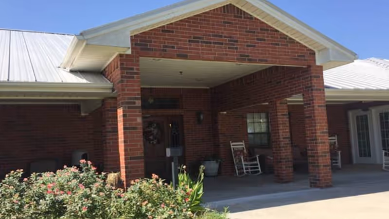 The brick retirement home front entrance features rocking chairs and shrubs.