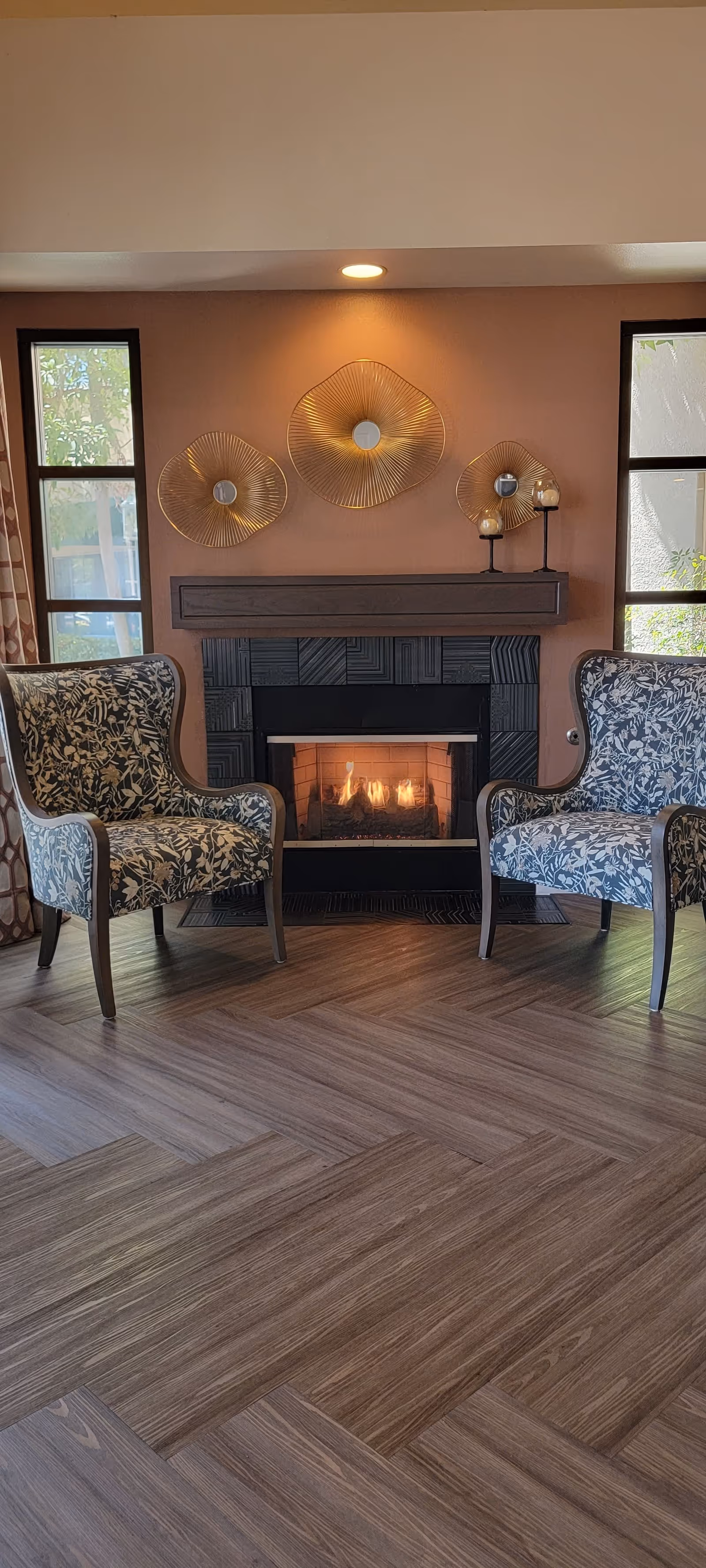 A cozy seating area with two patterned armchairs facing a lit fireplace. The fireplace has a dark geometric tile surround and a wooden mantel with decorative candle holders. Above the mantel, there are three gold wire wall art pieces. Two tall windows flank the fireplace, letting in natural light.