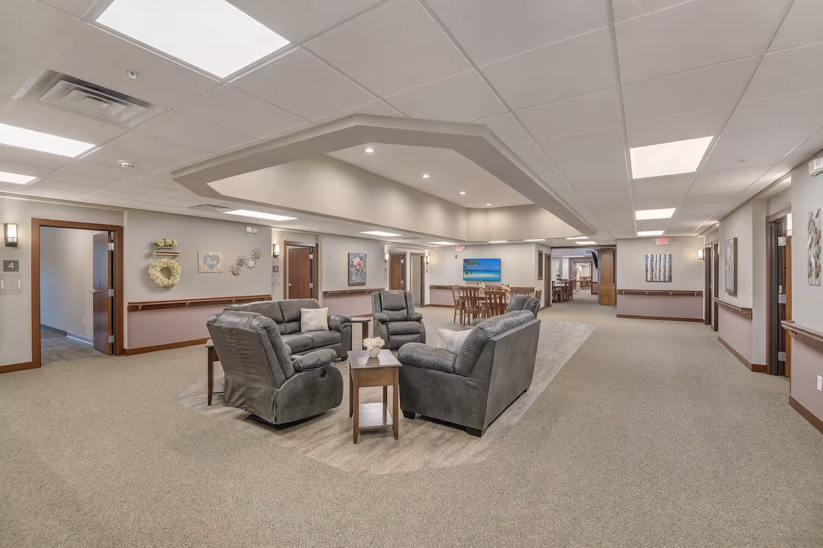 A spacious senior living facility common area with four gray recliner chairs arranged around a wooden coffee table with a small flower vase. The room has beige walls with wooden trim, several doors leading to other rooms, and a dining area with wooden tables and chairs in the background. The ceiling has recessed lighting and a decorative tray ceiling feature.