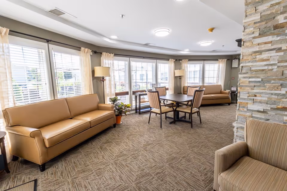 A bright and spacious common area with large windows covered by white blinds and light curtains. The room features two tan leather sofas, a small table with four striped chairs in the center, a striped armchair in the foreground, two floor lamps, and a stone accent wall on the right side. The carpet has a geometric pattern, and the ceiling has recessed lighting and round light fixtures.