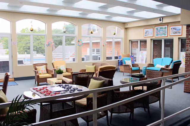 Sunlit communal lounge with wicker chairs and tables, colorful cushions, games, and large arched windows.
