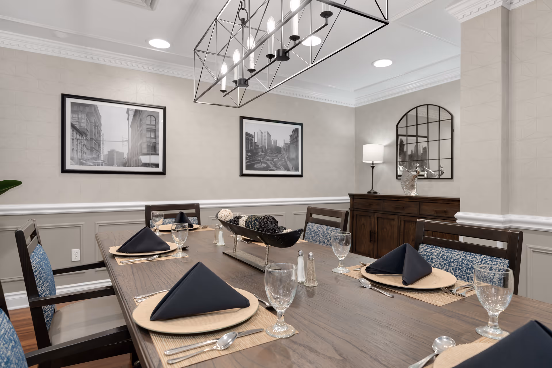A dining room with a wooden table set for six people, featuring black folded napkins, clear water glasses, and silverware on beige placemats. The room has beige walls with white wainscoting, two black and white framed cityscape photos on the wall, a dark wooden sideboard with a lamp and decorative items, and a large rectangular chandelier hanging above the table.