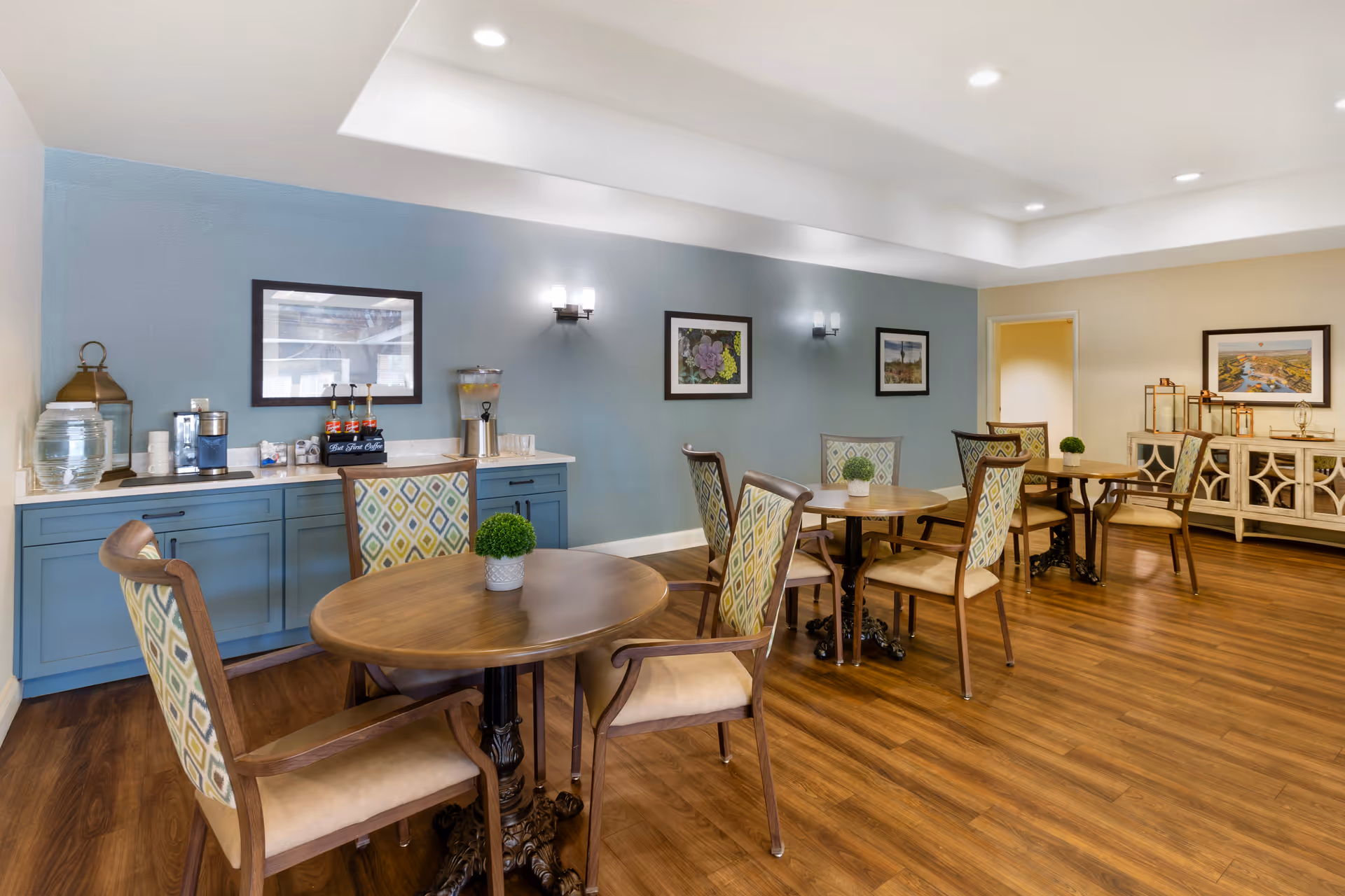 A bright and clean dining area with wooden floors, round wooden tables, and cushioned chairs with patterned backs. A blue sideboard against the wall holds a water dispenser, coffee machine, and cups. The walls are decorated with framed pictures and wall sconces providing soft lighting.