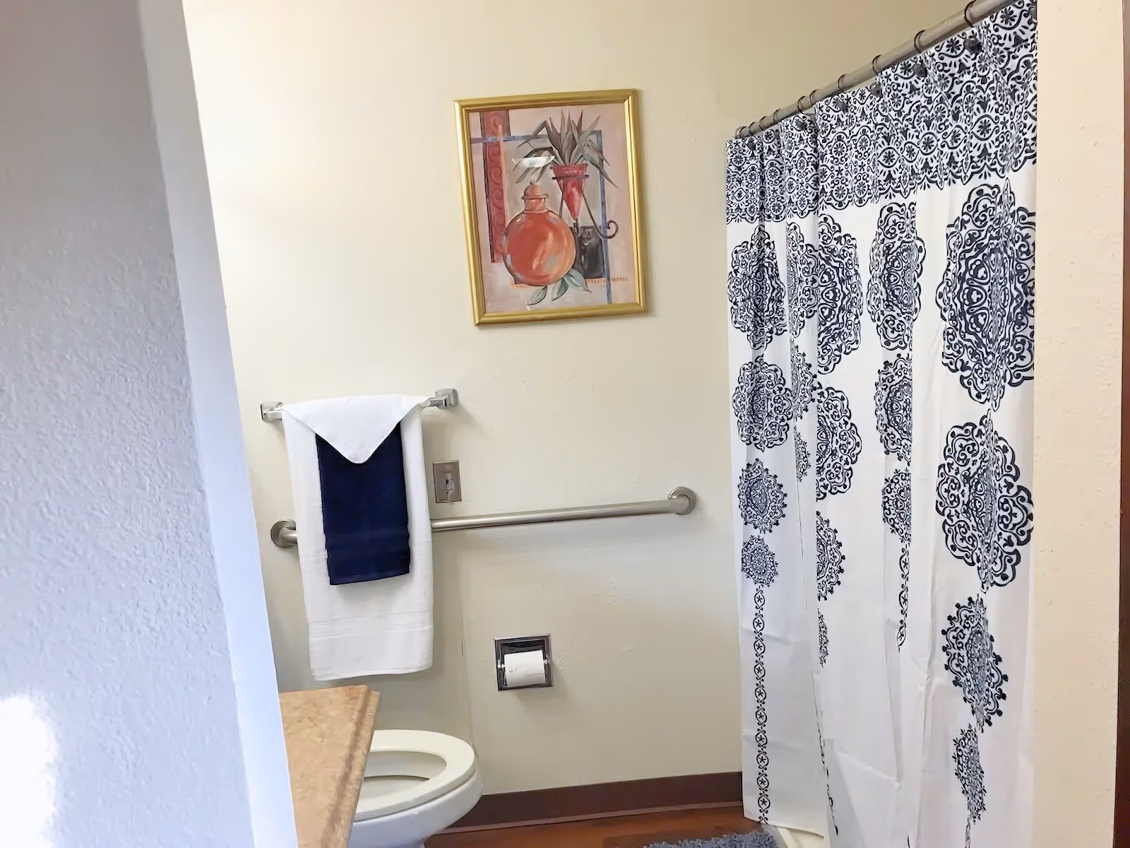 Small bathroom with a toilet, grab bar, towel on a rack, framed art, and a patterned shower curtain.