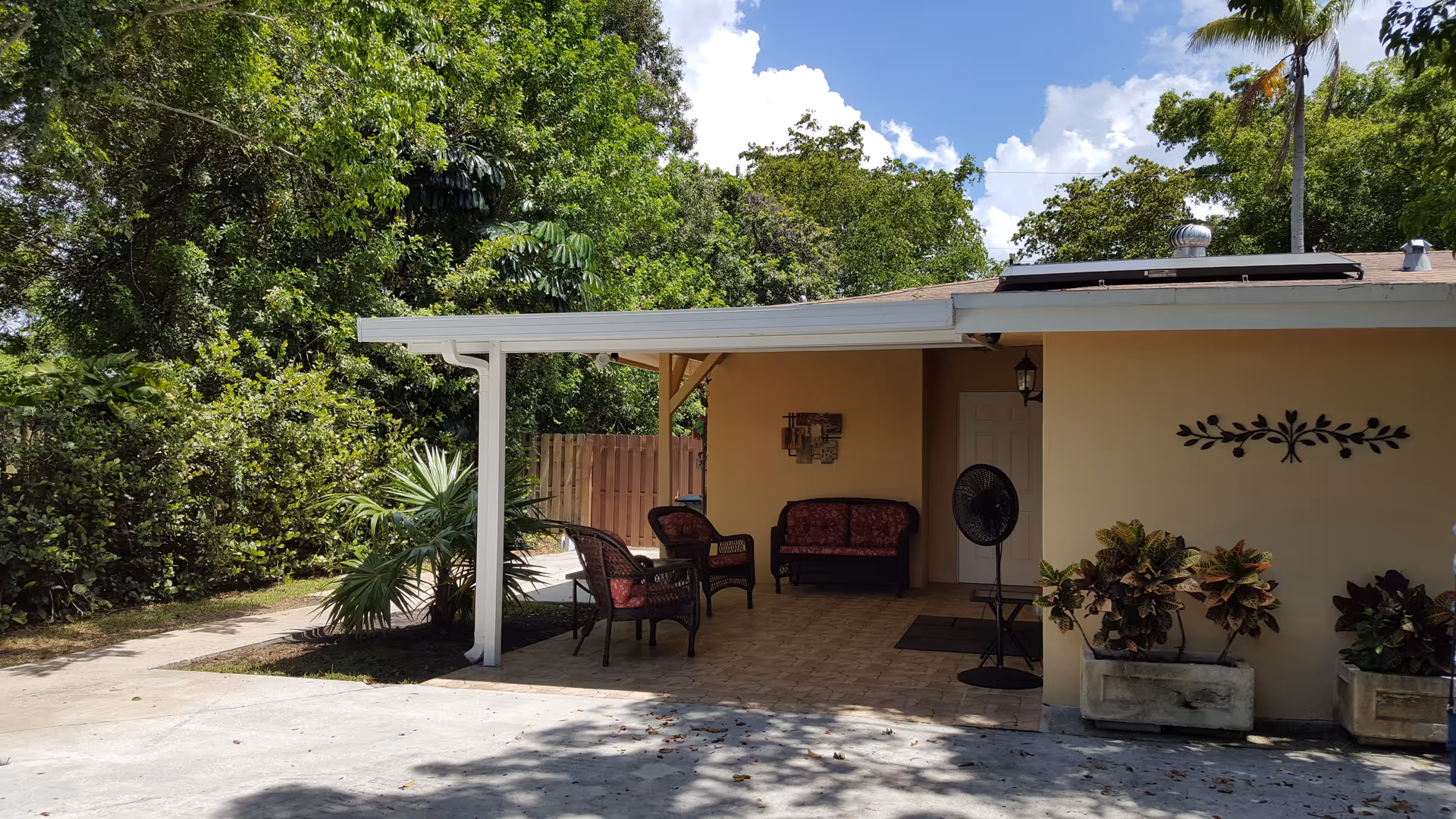 Outdoor covered patio area with seating including a loveseat and two chairs with red cushions. There is a standing fan and potted plants near the beige wall of the building. The patio is surrounded by greenery and trees under a partly cloudy blue sky.