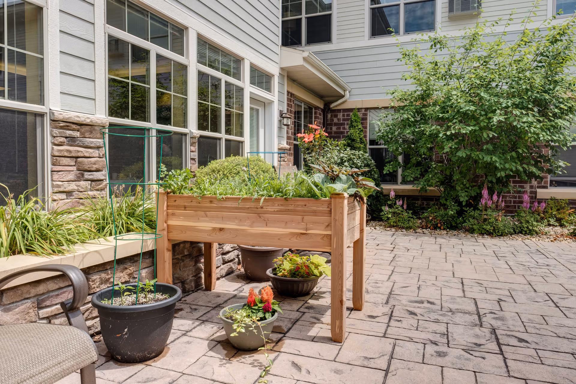 Outdoor patio area at Heritage Middleton featuring a raised wooden garden bed filled with various green plants and flowers. Several potted plants are placed on the stone-paved ground, and the building exterior includes large windows, stone and brick walls, and siding. There is a chair partially visible on the left side.
