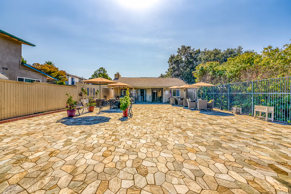 Outdoor patio area with stone tile flooring, several seating arrangements including benches and chairs with umbrellas, potted plants, and a building in the background under a clear blue sky.