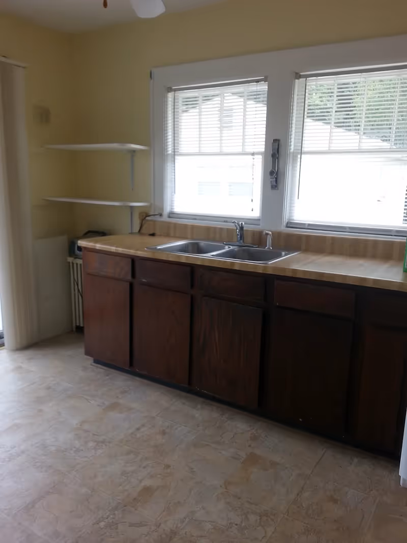 A kitchen area with dark wooden cabinets, a double stainless steel sink, and a beige countertop. Two windows with white blinds are above the sink, letting in natural light. There are two white wall-mounted shelves on the left side and a beige tiled floor.
