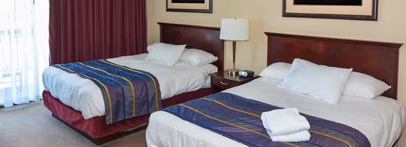Two neatly made double beds with white linens and blue-striped runners in a bright bedroom with a nightstand and lamp between them.
