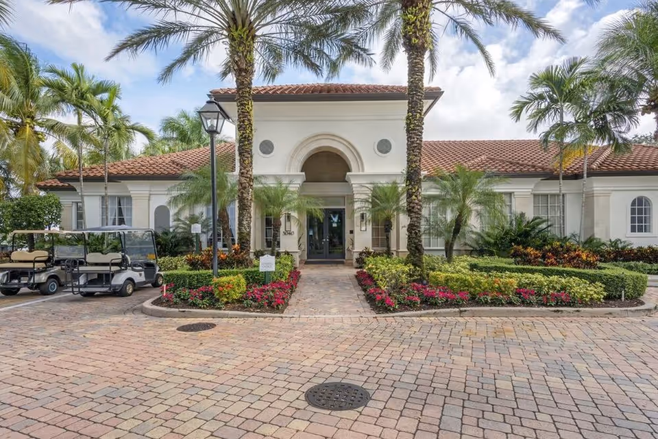 Front entrance of a Mediterranean-style building with palm trees, landscaped flowerbeds and parked golf carts in the driveway.