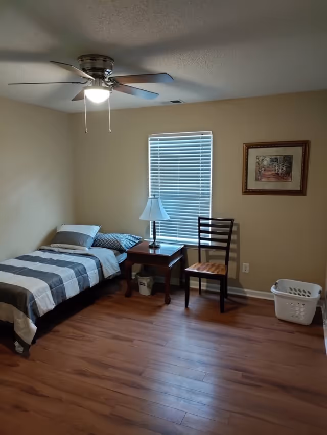 Small bedroom with a single bed, nightstand and lamp, chair, window with blinds and a ceiling fan.