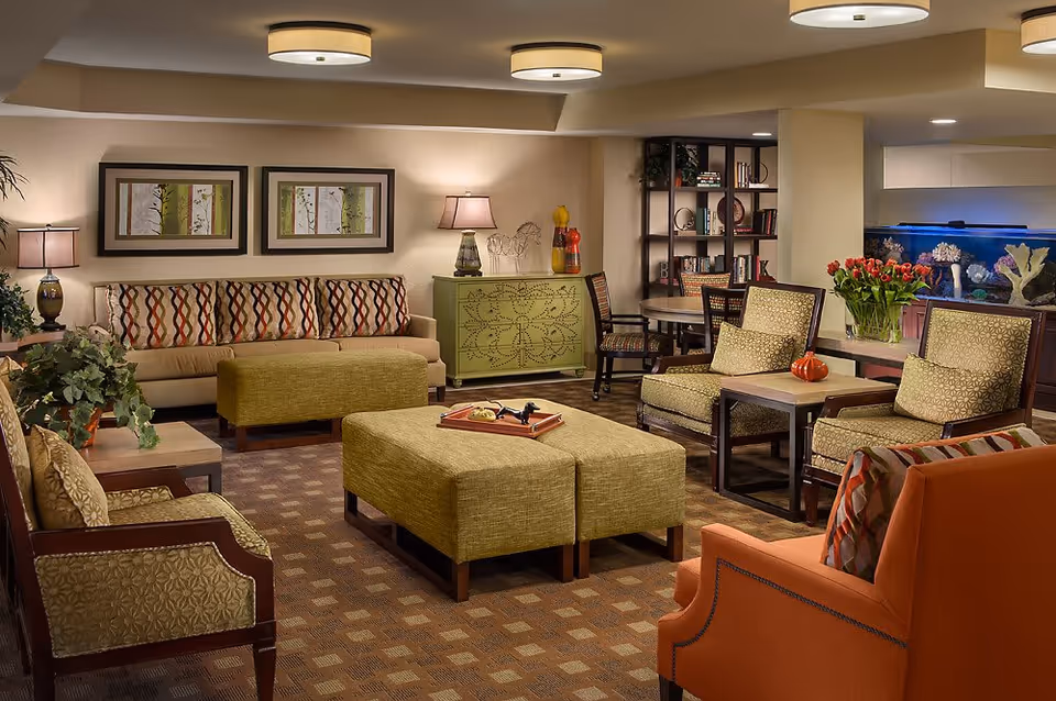 A cozy senior living facility lounge with a beige sofa adorned with patterned cushions, two green armchairs, an orange armchair, and a large green ottoman in the center. The room features warm lighting from ceiling fixtures and table lamps, framed artwork on the wall, a green cabinet, a bookshelf, a round table with chairs, and a large aquarium with coral decorations. There are also plants and a vase of red tulips adding a touch of nature to the space.