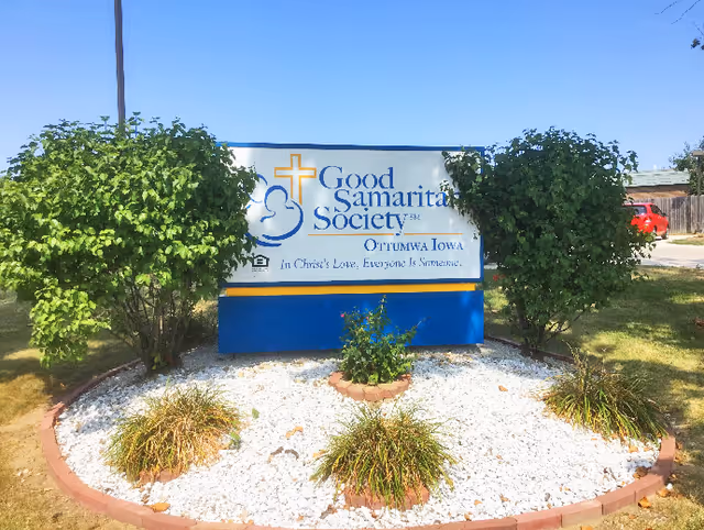 Outdoor sign for Good Samaritan Society in Ottumwa, Iowa, surrounded by bushes and landscaping with white rocks and small plants under a clear blue sky.