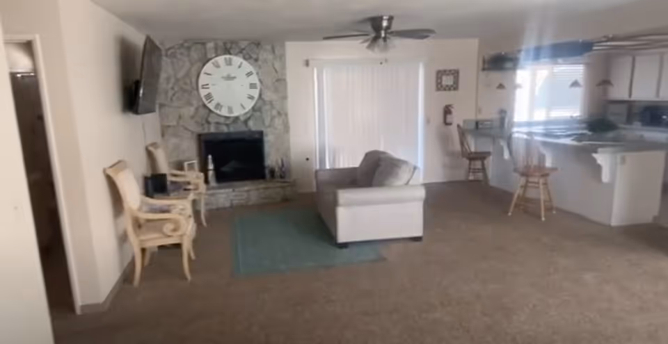 Open living area with a stone fireplace, sofa and chairs facing a wall-mounted TV and an adjacent kitchen with bar stools.
