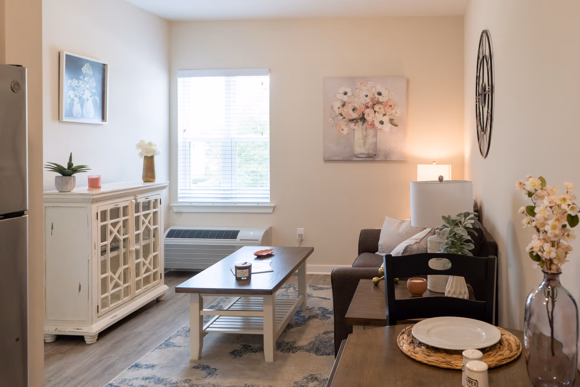 A cozy living room area with a brown sofa, a wooden coffee table, a side table with a lamp and plant, a white cabinet, and a dining table set with a plate and salt and pepper shakers. The room has light-colored walls decorated with floral paintings and a large clock, and a window with blinds letting in natural light.