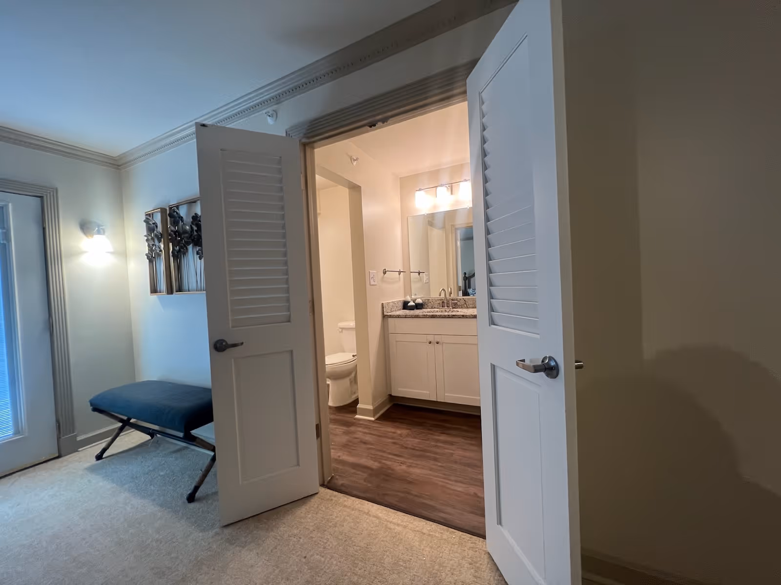 View of a bathroom through partially open double doors from an adjacent room. The bathroom features a white vanity with a granite countertop, a large mirror, and a toilet. The adjacent room has a cushioned bench and wall-mounted decorative art, with a wall light fixture and a door with blinds.