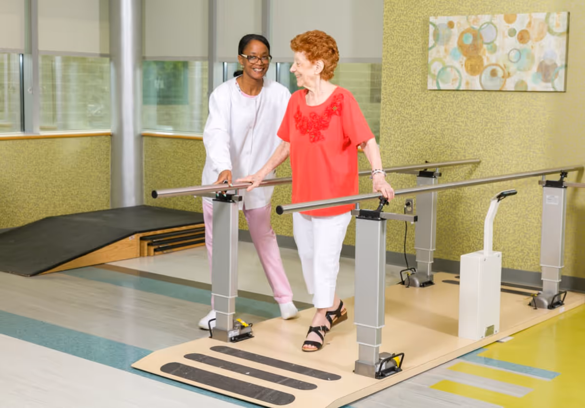 A healthcare worker assists an elderly woman as she walks between parallel bars in a rehabilitation or physical therapy room. The elderly woman is wearing a red top and white pants, and the healthcare worker is dressed in white and pink scrubs. The room has large windows, a ramp, and a piece of abstract wall art.