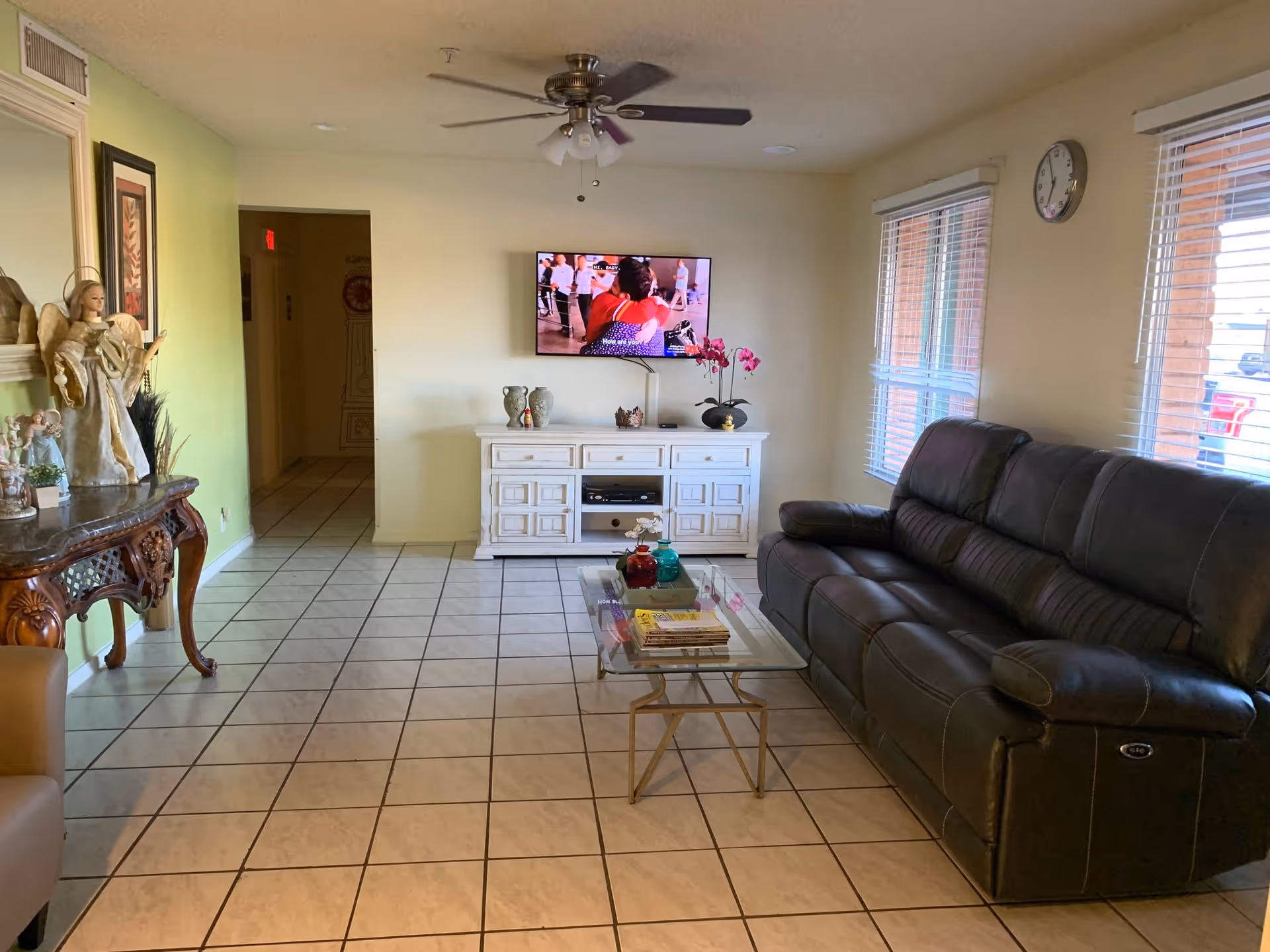 A cozy living room in a senior living facility featuring a black leather sofa, a glass coffee table with books and decorative items, a white TV stand with vases and a potted orchid, a wall-mounted flat-screen TV, two windows with blinds, a ceiling fan with lights, and a decorative wooden console table with angel figurines and framed artwork on the left wall.