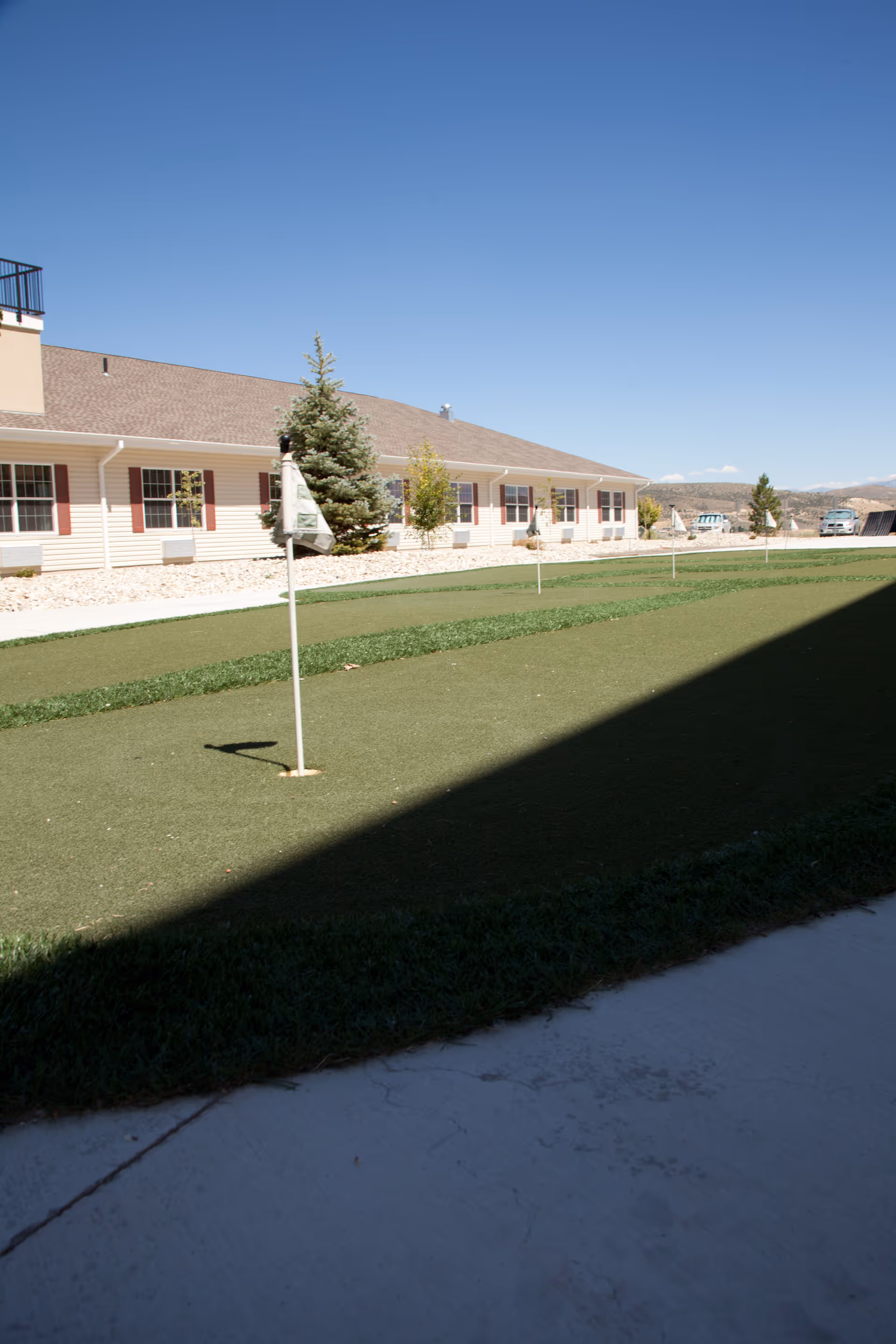 Outdoor putting green with flags in front of a single-story residential building under a clear blue sky.