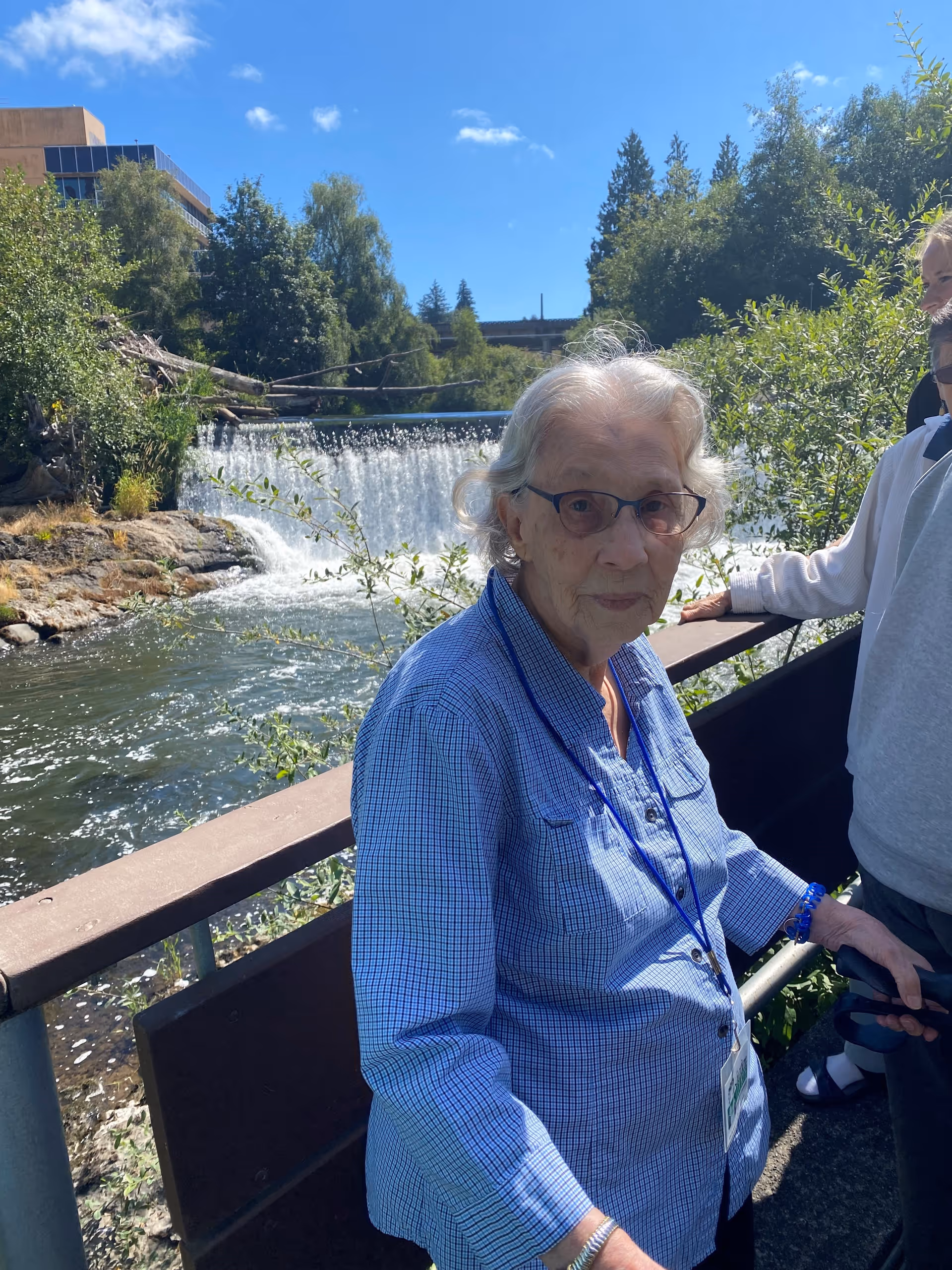 An elderly woman wearing glasses and a blue checkered shirt stands outdoors near a railing with a waterfall and lush greenery in the background. Another person is partially visible beside her.