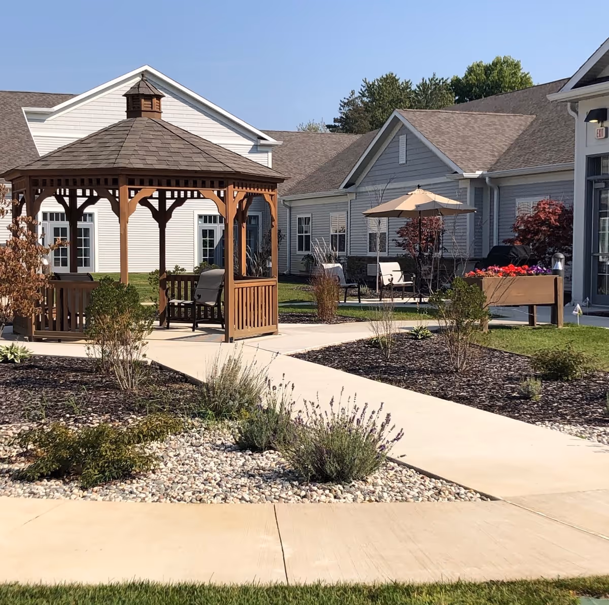 Outdoor garden area at North Woods Village of Kalamazoo featuring a wooden gazebo with seating, a paved walkway, landscaped plants, and patio furniture with an umbrella near a building.