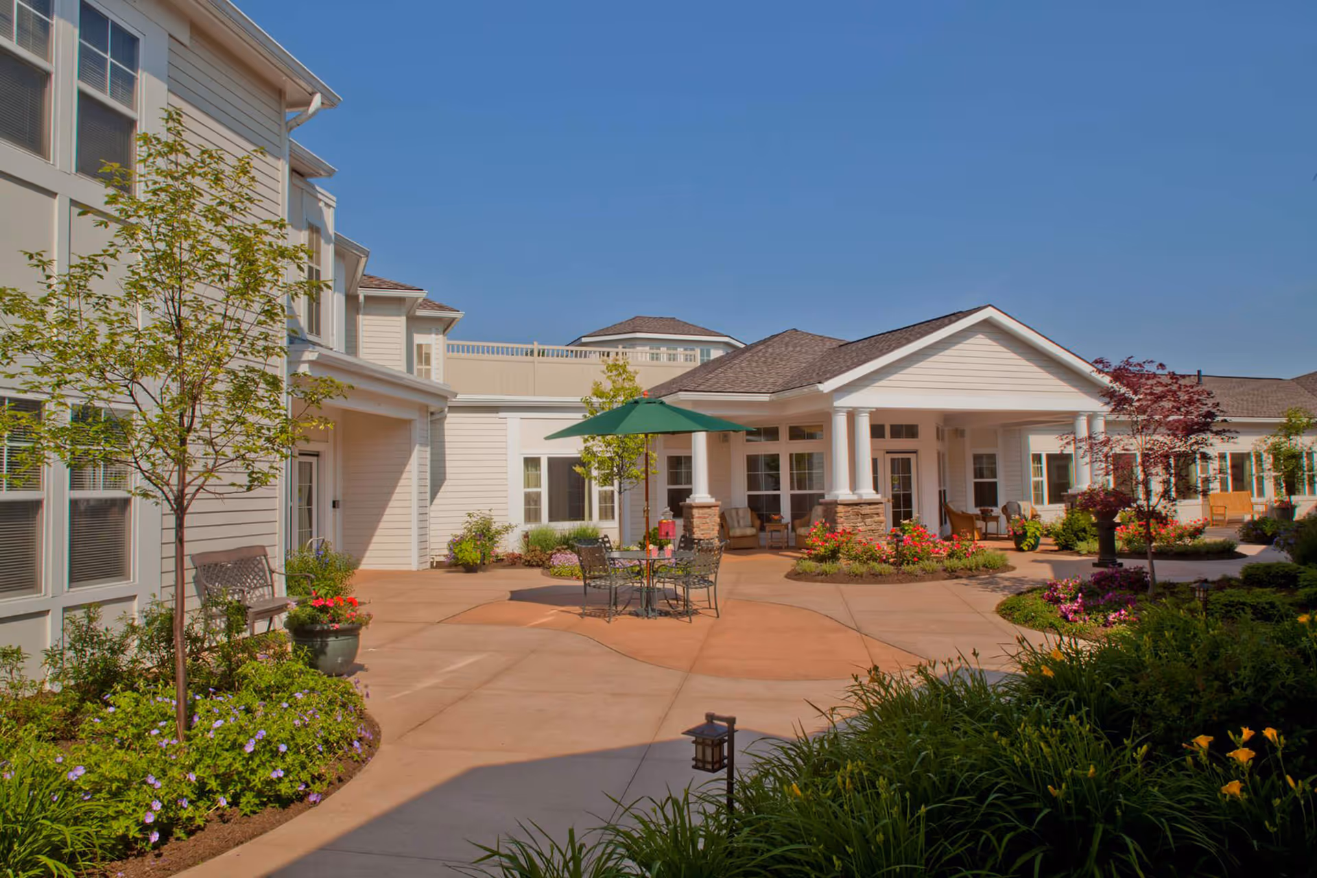 Outdoor courtyard area of Glenmere Assisted Living featuring a paved patio with a table, chairs, and a green umbrella. The surrounding building is light-colored with white trim and columns. There are landscaped garden beds with various plants and flowers, small trees, and benches along the building walls under a clear blue sky.