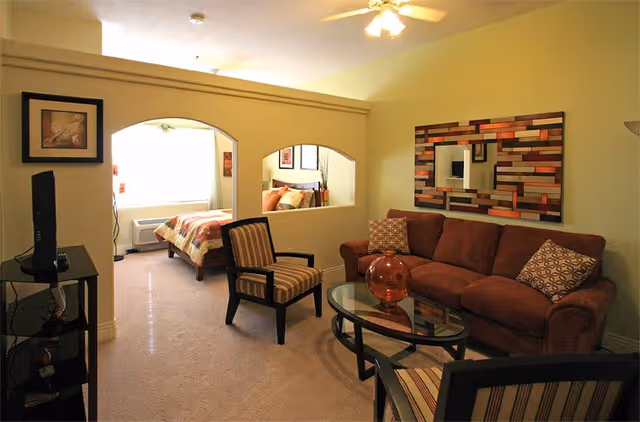 A cozy living room area with a brown sofa, two striped armchairs, and a glass coffee table with a decorative orange vase. The room has a ceiling fan with lights and a colorful rectangular wall mirror. Through two arched openings in the wall, a bedroom with a bed and framed artwork is visible.