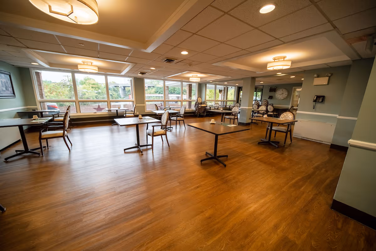 A spacious dining room with wooden floors and multiple tables and chairs arranged throughout. Large windows line one wall, allowing natural light to fill the room. Ceiling lights provide additional illumination. The room appears clean and well-maintained, with a clock and a telephone mounted on one wall.