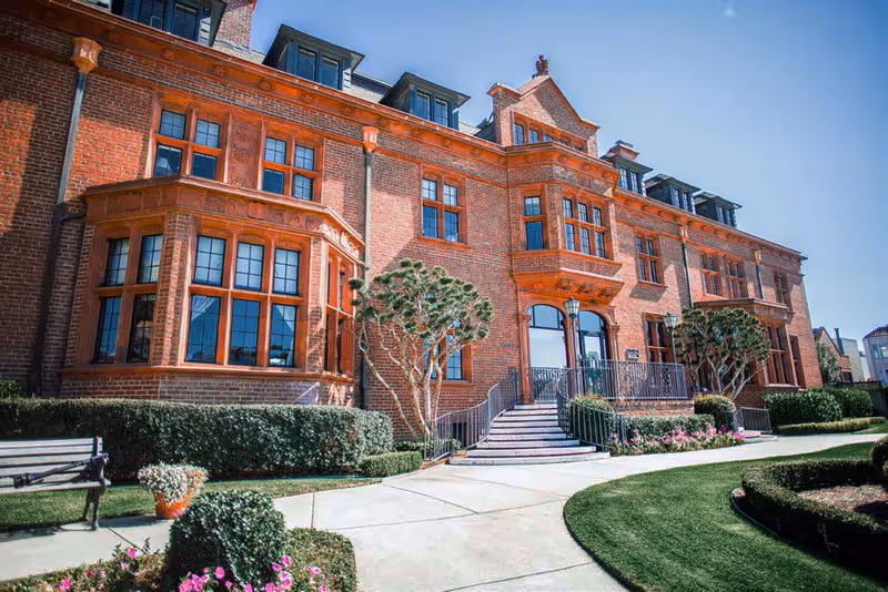 Red brick, multi-story building front with large bay windows, entrance steps, and landscaped walkway.