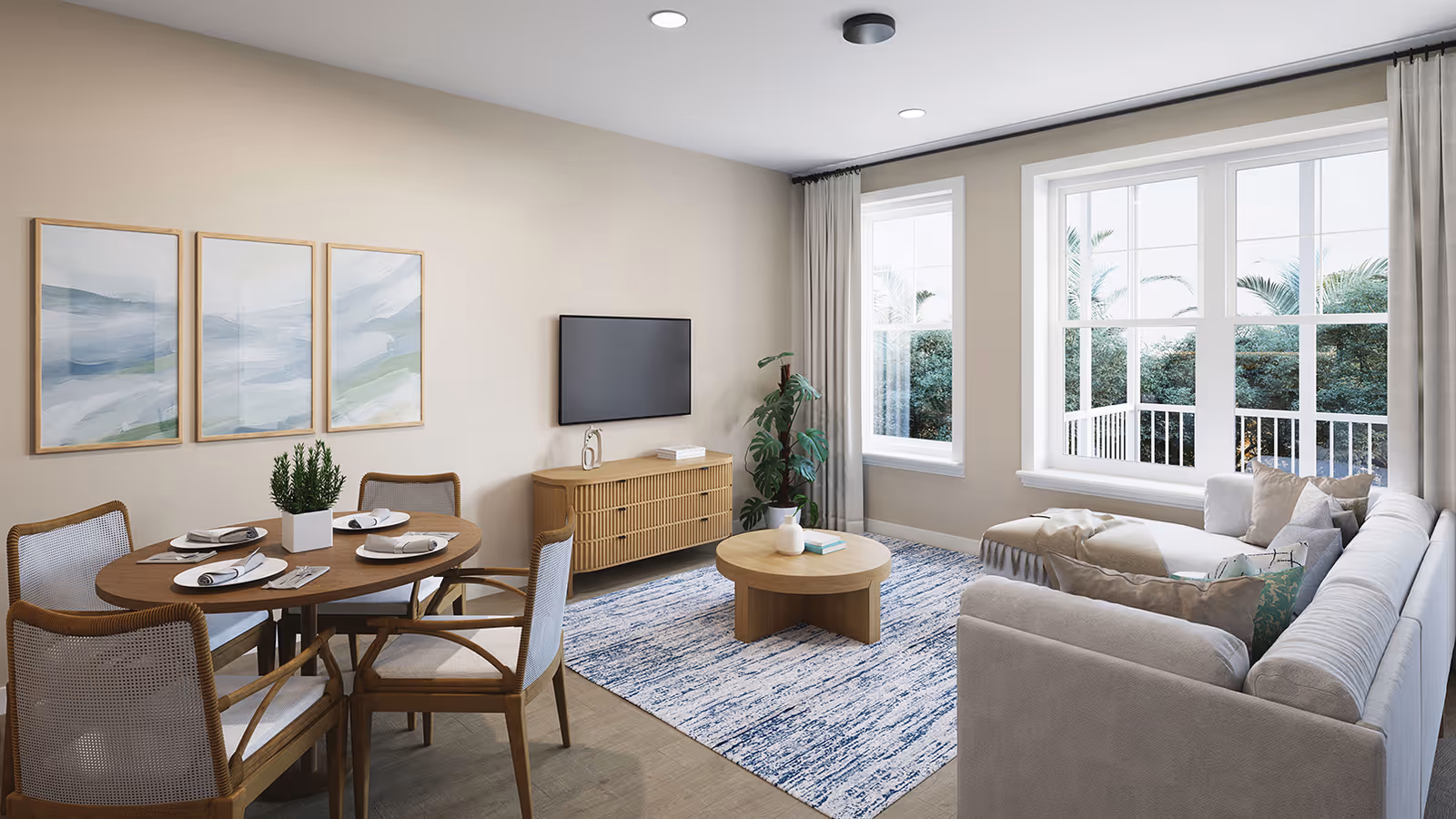 A bright and modern living room and dining area in a senior living community. The room features a round wooden dining table set with four chairs, each with light-colored cushions. On the wall above the dining table are three framed abstract paintings in soft blue and green tones. A flat-screen TV is mounted on the wall above a wooden media console. A large window with white curtains lets in natural light and shows greenery outside. A light-colored sectional sofa with multiple cushions faces a round wooden coffee table on a blue and white patterned rug. A potted plant is placed near the window.
