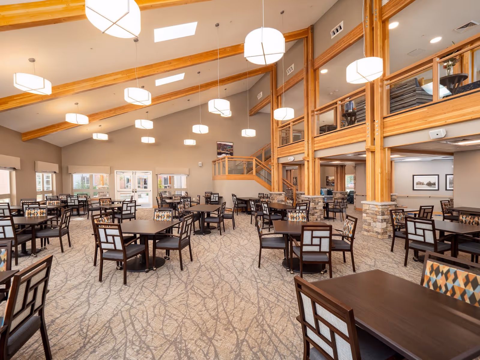 Spacious dining room in a senior living community with multiple dark wood tables and chairs arranged neatly on a patterned carpet. The room features high ceilings with wooden beams, large windows allowing natural light, and modern hanging light fixtures. There is a staircase and a mezzanine area with glass railings on one side.