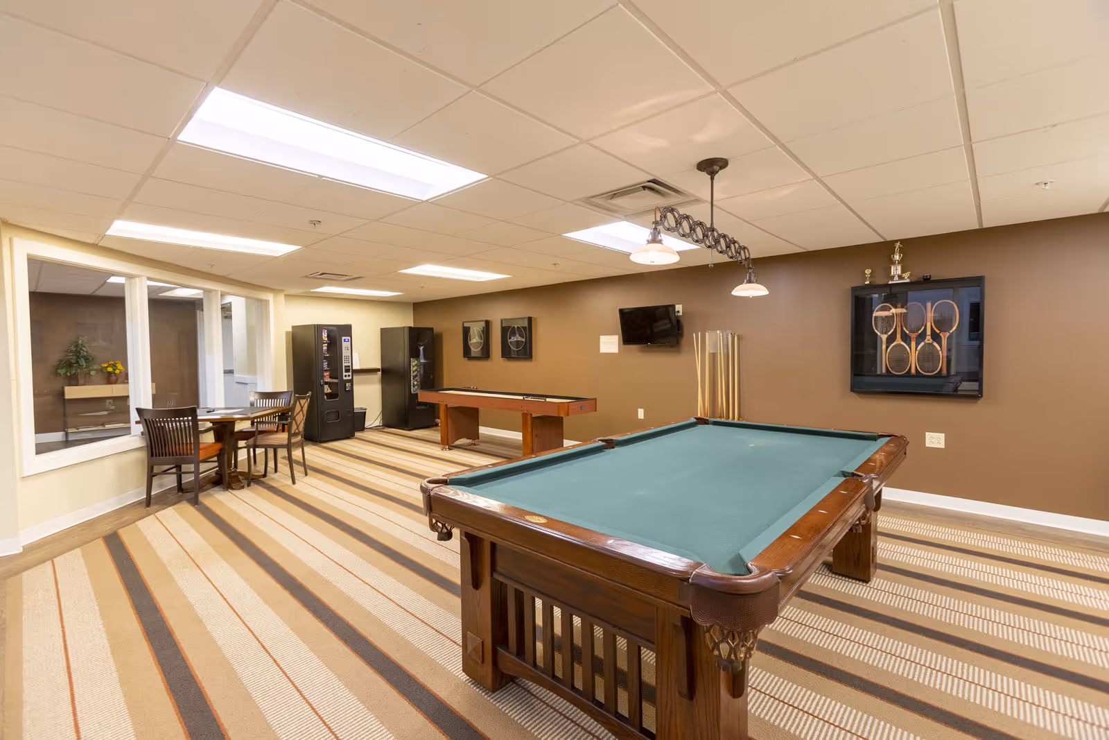 A recreational room with a pool table in the foreground and a shuffleboard table against a brown wall. The room has striped carpet flooring, a small table with four chairs near a window, two vending machines, and a wall-mounted TV. There are also framed tennis rackets and trophies displayed on the wall.