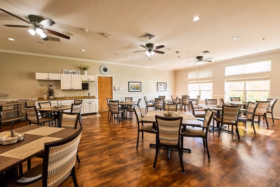 Bright communal dining room with multiple tables and chairs, ceiling fans, large windows, and a small kitchenette along the back wall.