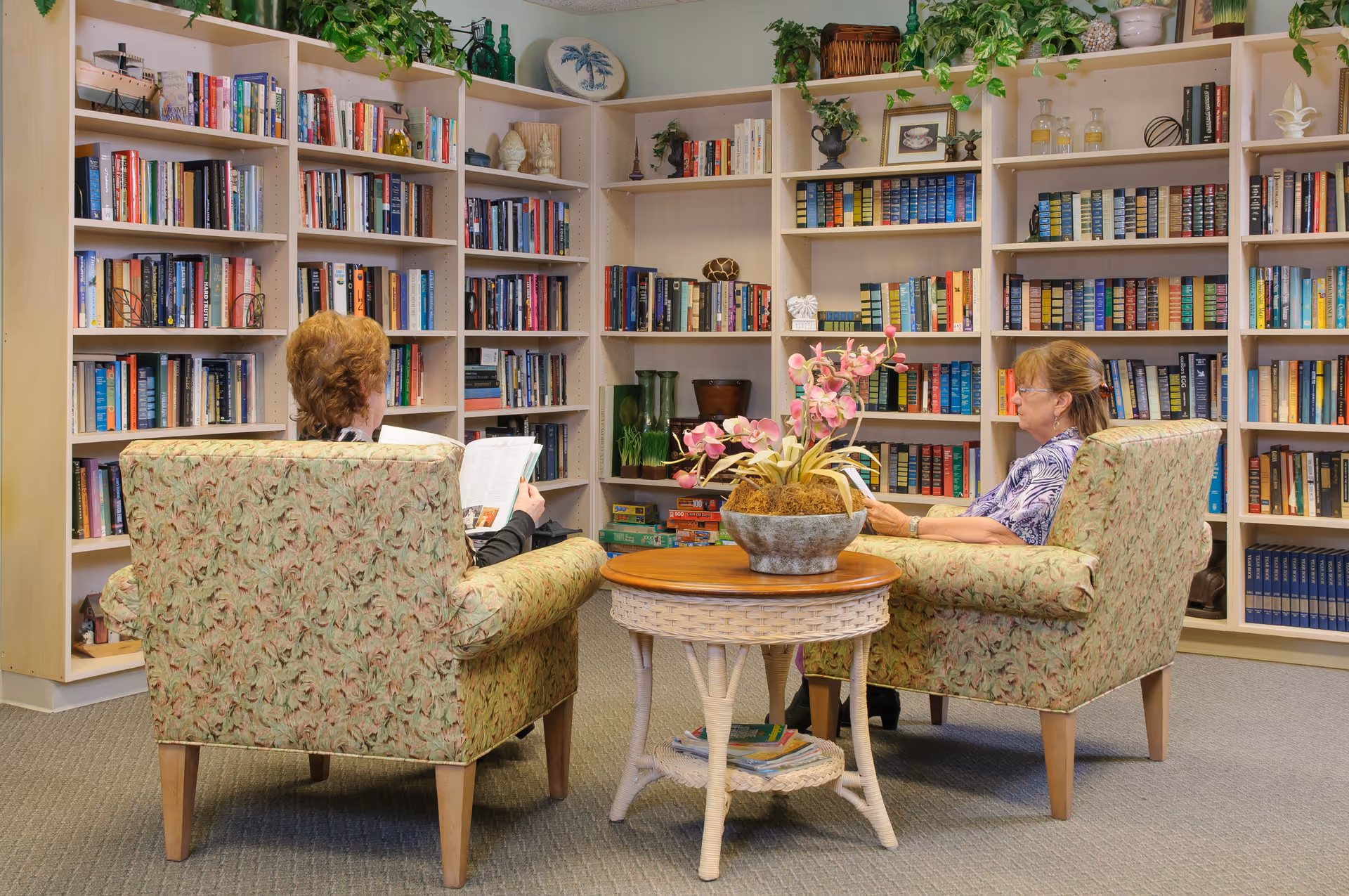 Two elderly women sitting in floral armchairs facing bookshelves filled with books in a cozy library or reading room. A round wicker table with a flower arrangement is placed between them.