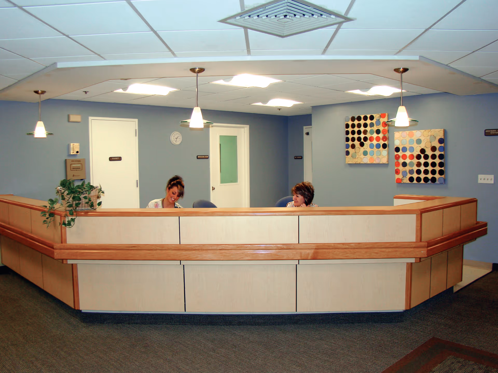 Reception desk in a senior living facility lobby with two staff seated behind it, pendant lights, blue walls and abstract artwork.