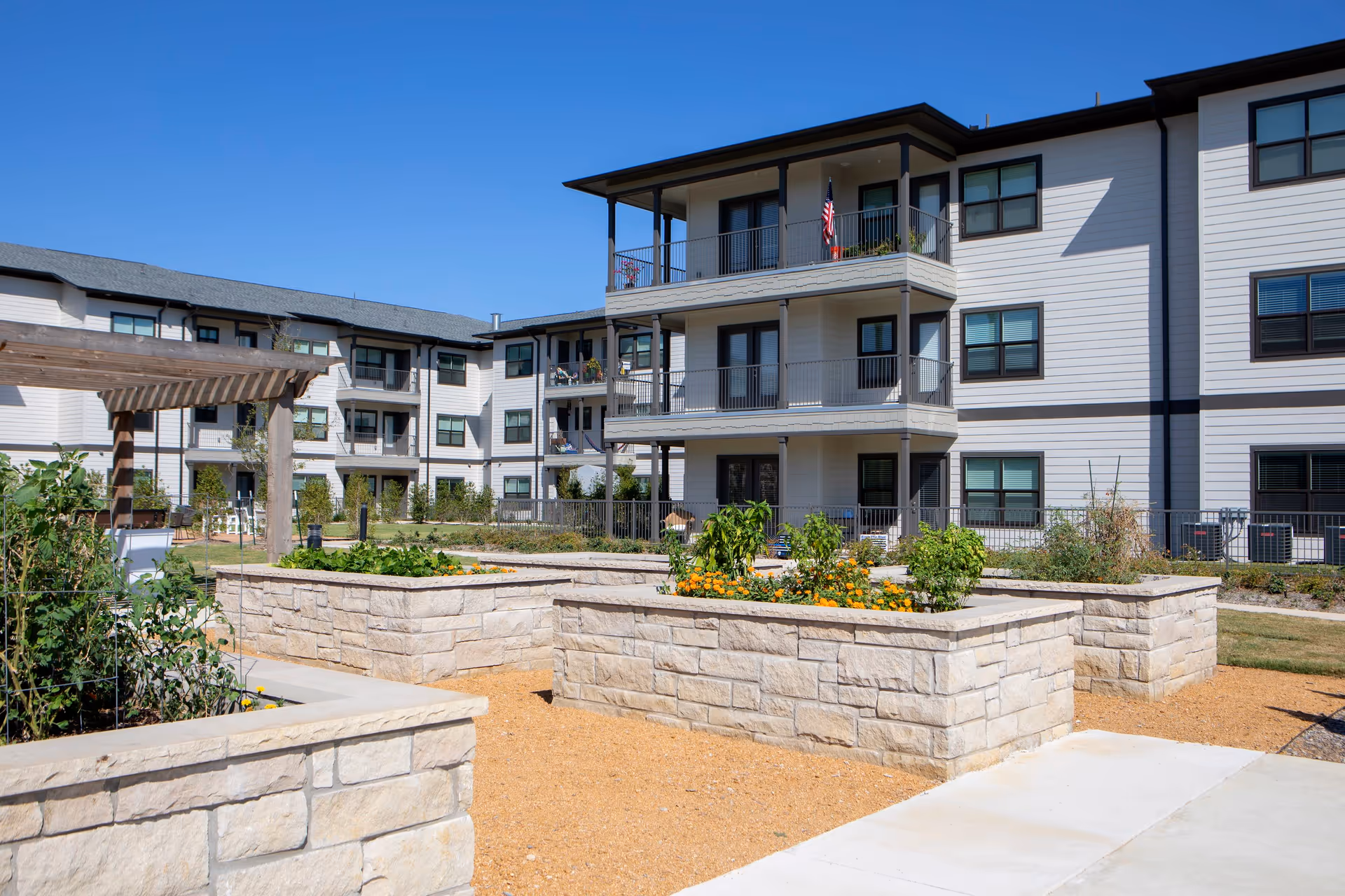 Outdoor garden area with raised stone flower beds containing plants and flowers, situated in front of a three-story residential building with balconies and windows under a clear blue sky.
