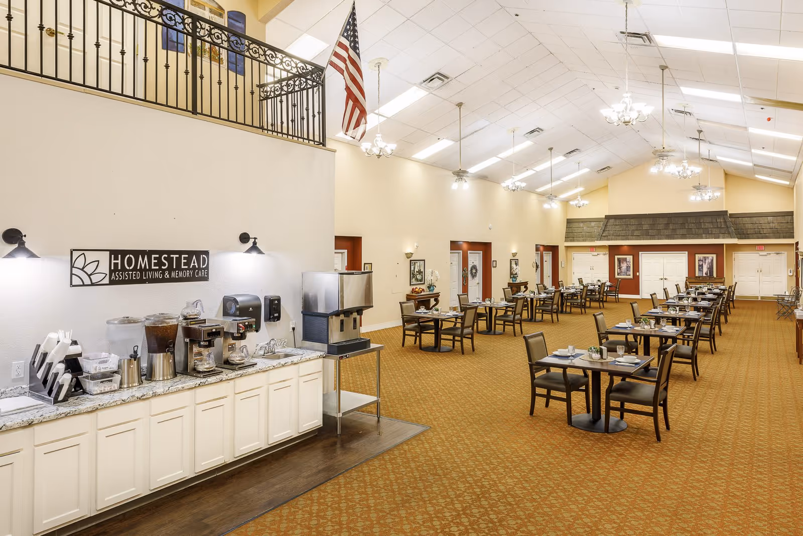 A spacious dining room in an assisted living facility with multiple tables and chairs arranged neatly. On the left side, there is a beverage station with coffee machines, water dispensers, and cups. The room has high ceilings with hanging chandeliers and an American flag hanging from the upper railing. The walls are decorated with framed pictures and the carpet has a patterned design.
