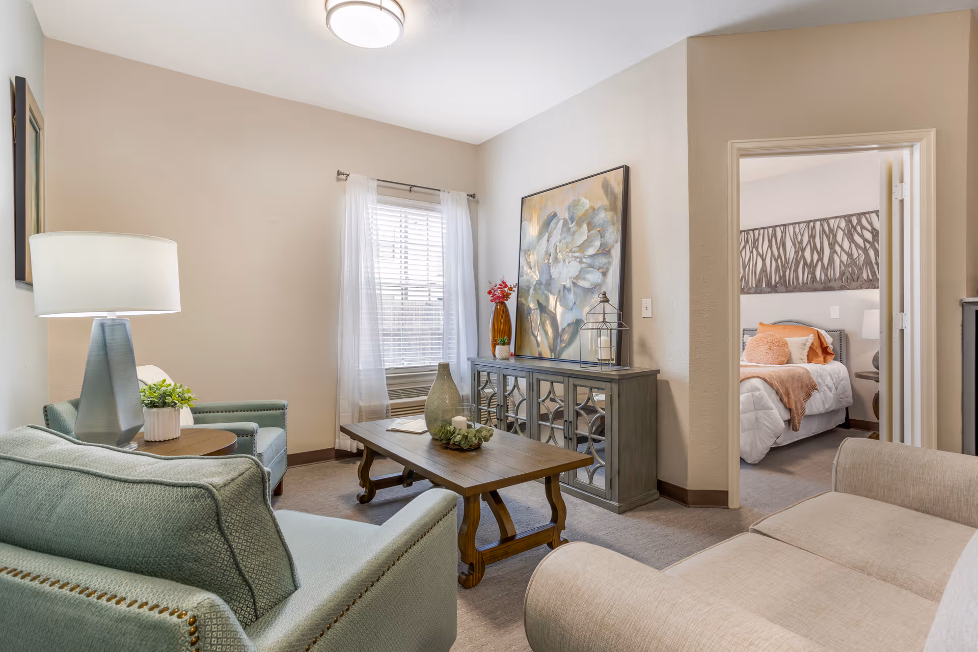 Cozy furnished living room with armchairs, a coffee table, a decorative console with artwork, and a view into a bedroom.
