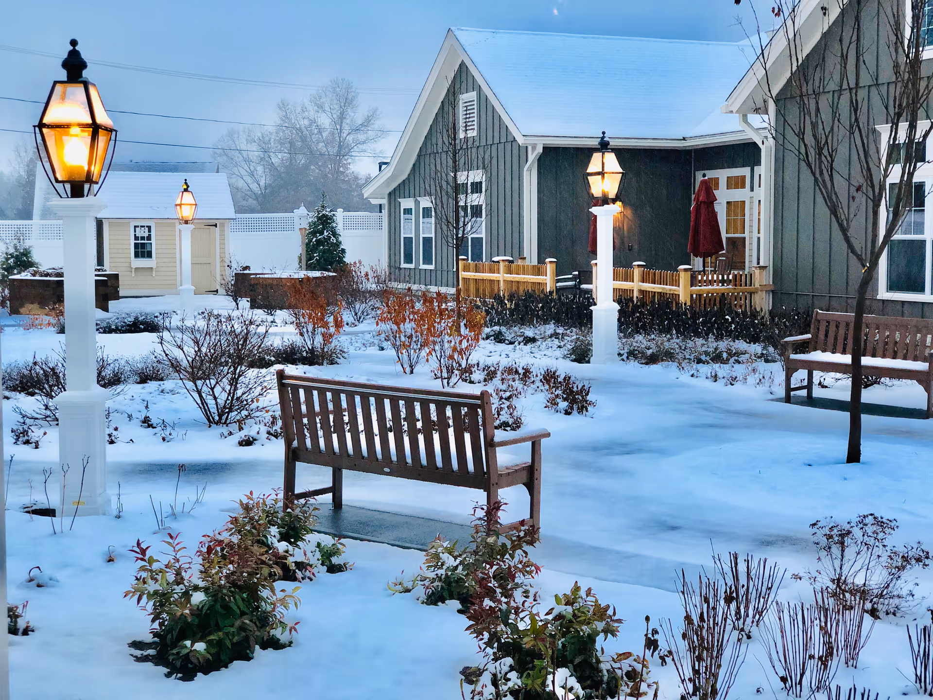 Snow-covered courtyard with wooden benches, lit lampposts, and a residential building in the background.