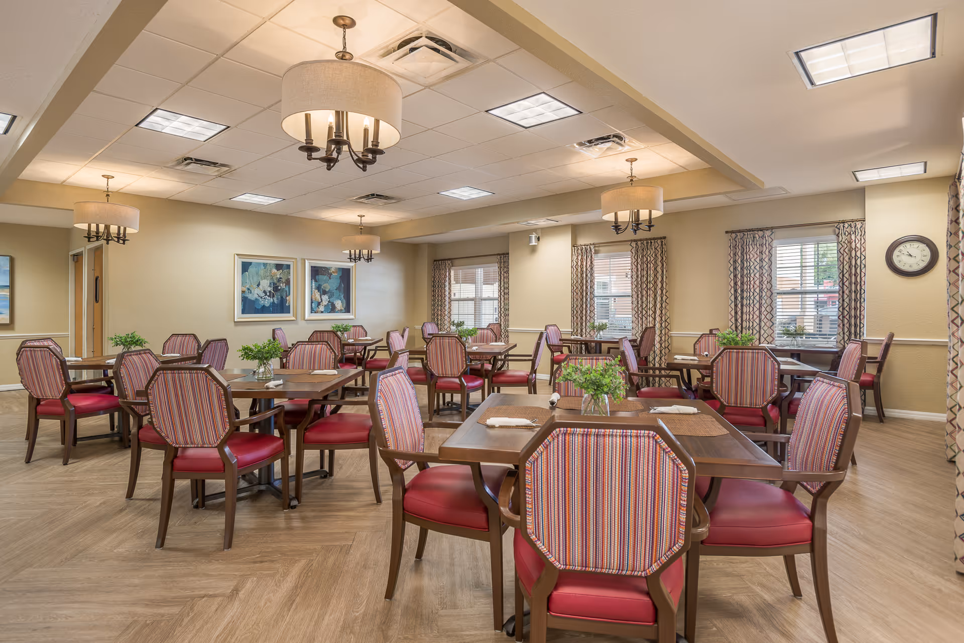 A bright dining room with multiple wooden tables and striped red-upholstered chairs, chandeliers, large windows, and neutral decor.