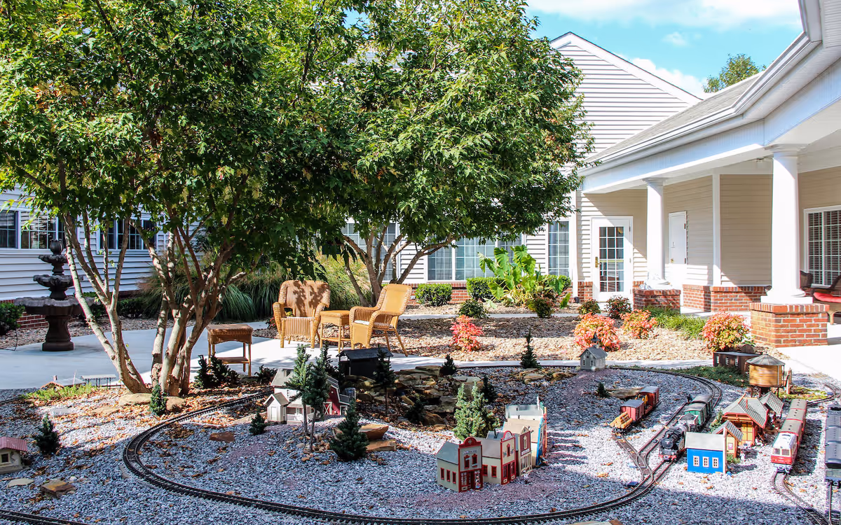 Outdoor courtyard area with a miniature train set on gravel surrounded by small model buildings and trees. There are wicker chairs and a table under leafy trees, with a beige building featuring white columns and windows in the background.