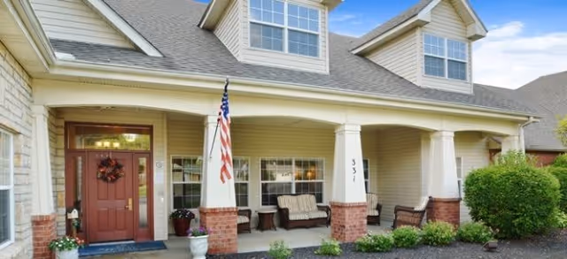 Front exterior view of a two-story residential building with a covered porch featuring seating, an American flag, and a red front door with a wreath. The building has beige siding, brick accents, and multiple windows.