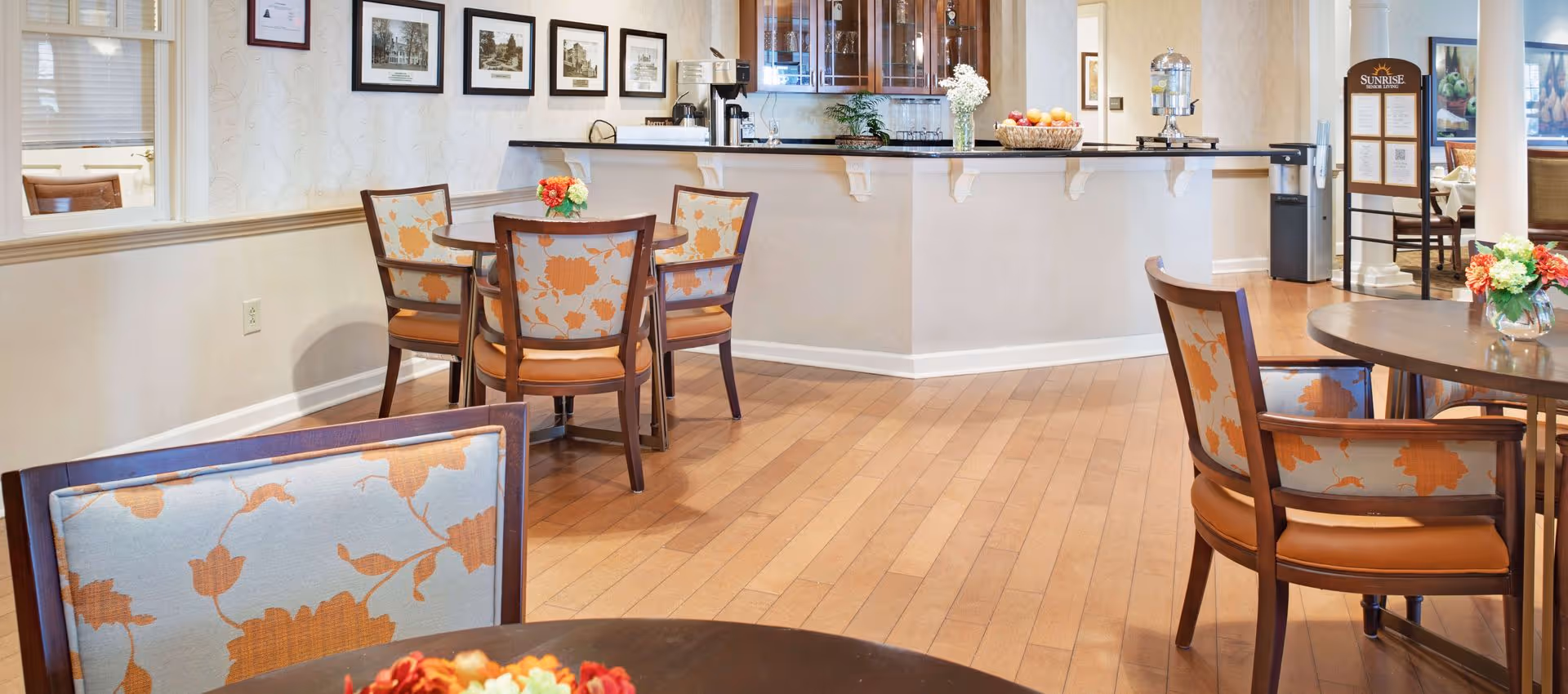 A bright and clean dining area in a senior living facility with wooden floors, round tables, and chairs upholstered with a floral pattern. There is a counter with a coffee machine, glass cabinets, and a water dispenser. A signboard with the Sunrise Senior Living logo is visible in the background.