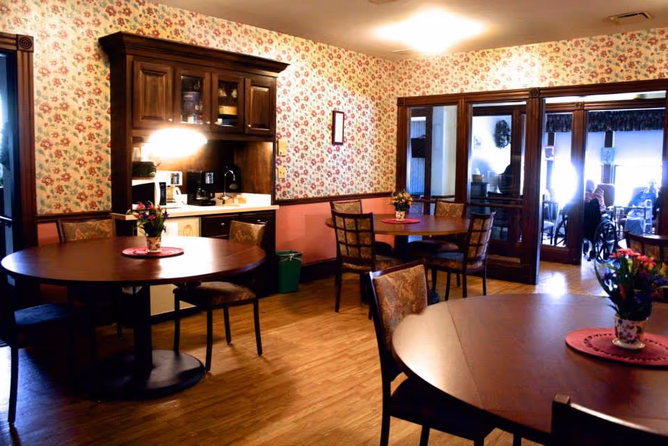 A cozy dining room with round wooden tables and cushioned chairs. Each table has a small floral centerpiece on a red doily. The walls are decorated with floral wallpaper and a wooden cabinet with a coffee maker and microwave is against one wall. The room has wooden flooring and glass doors leading to another room where people are seated.