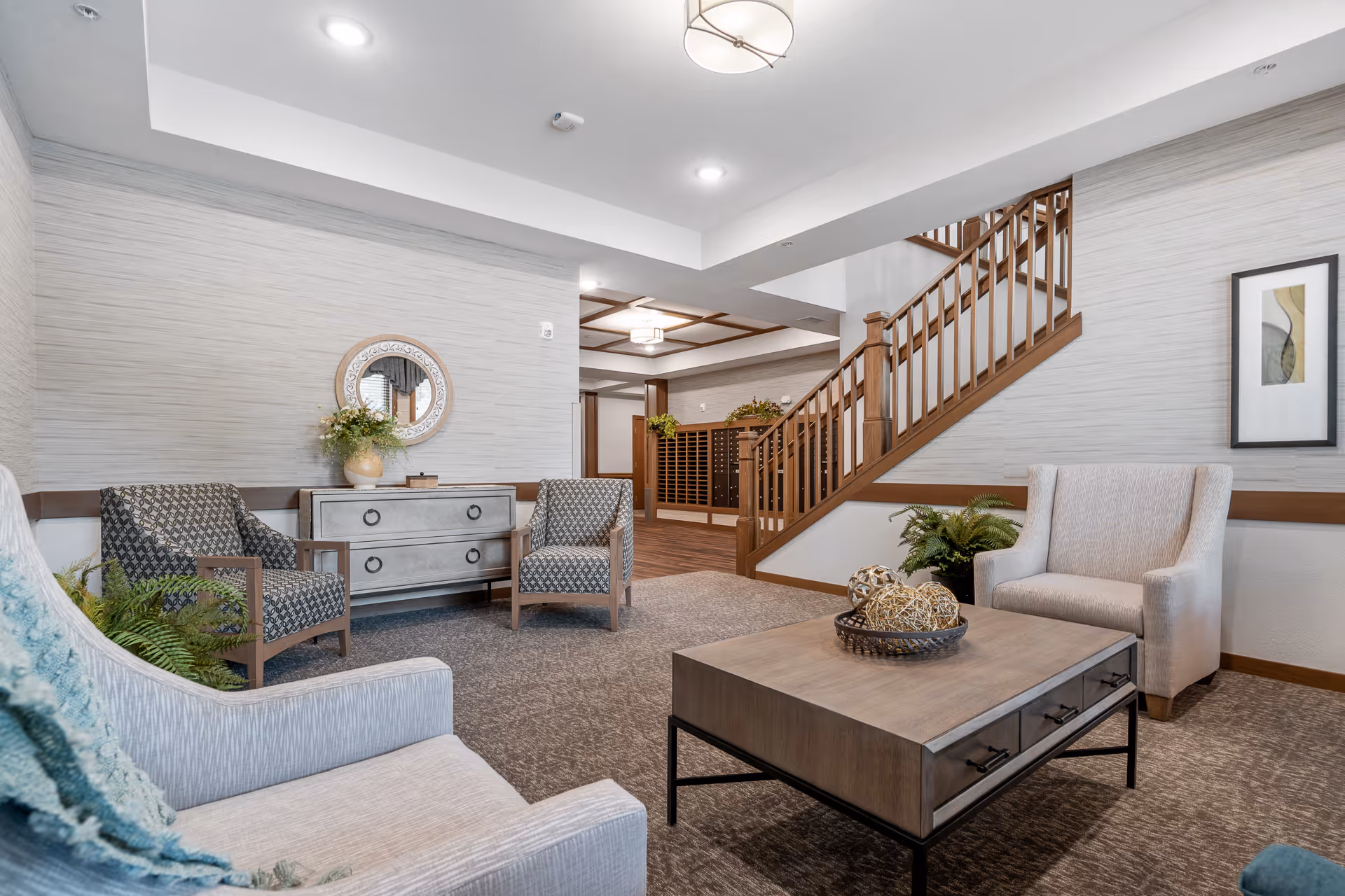A cozy seating area inside a senior living facility featuring two patterned armchairs, two beige armchairs, a wooden coffee table with decorative items, a small dresser with a round mirror above it, and a staircase with wooden railings. The walls are light-colored with wood trim, and there is a framed abstract artwork on one wall.