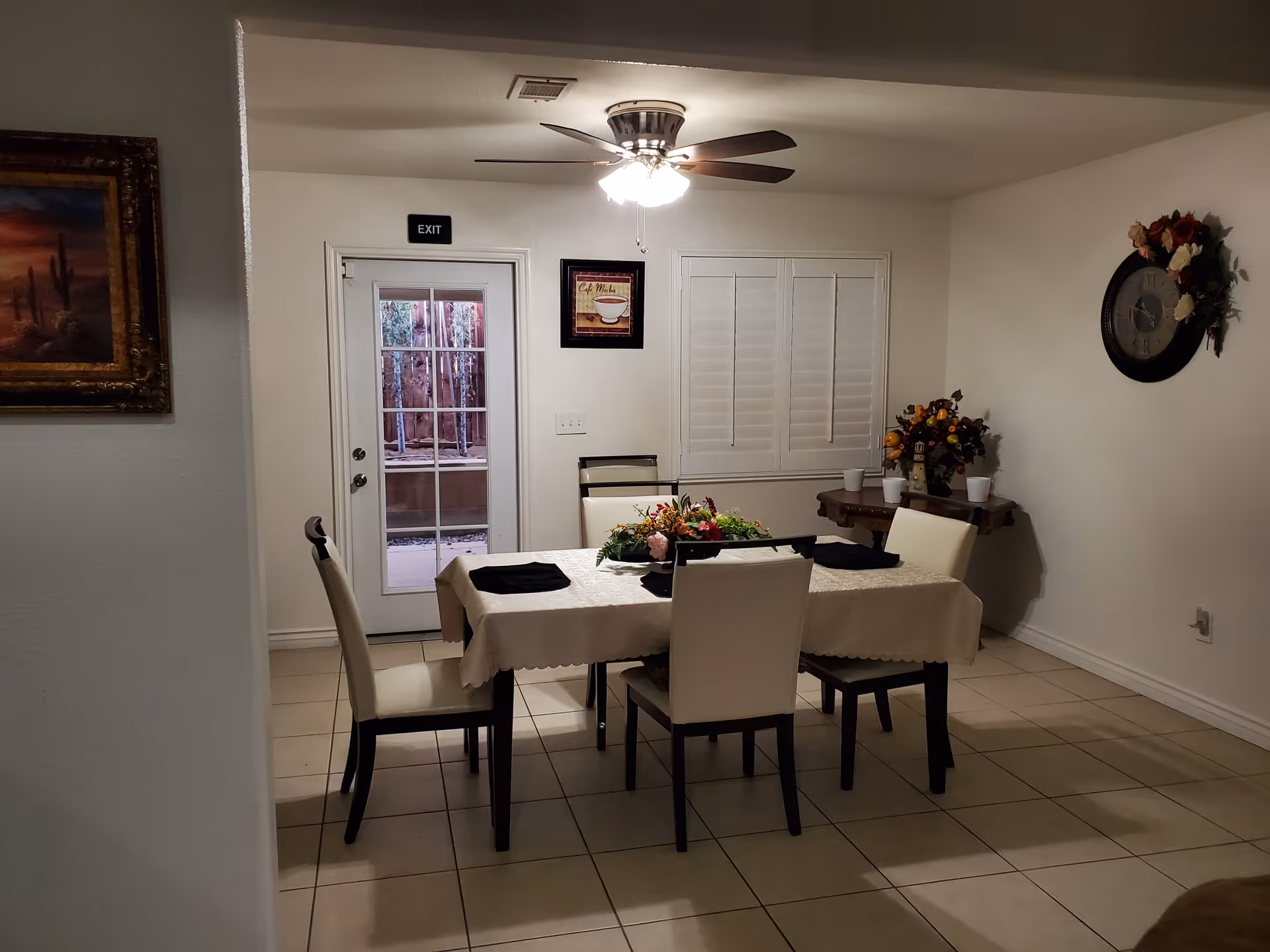 A dining room with a rectangular table covered with a white tablecloth and set with black napkins. Four white chairs with dark legs surround the table. A ceiling fan with lights is above the table. There is a glass door with an exit sign above it, a window with white shutters, a wall clock decorated with flowers, and a small table with a flower arrangement and cups in the corner. A framed painting of a desert scene is on the left wall.