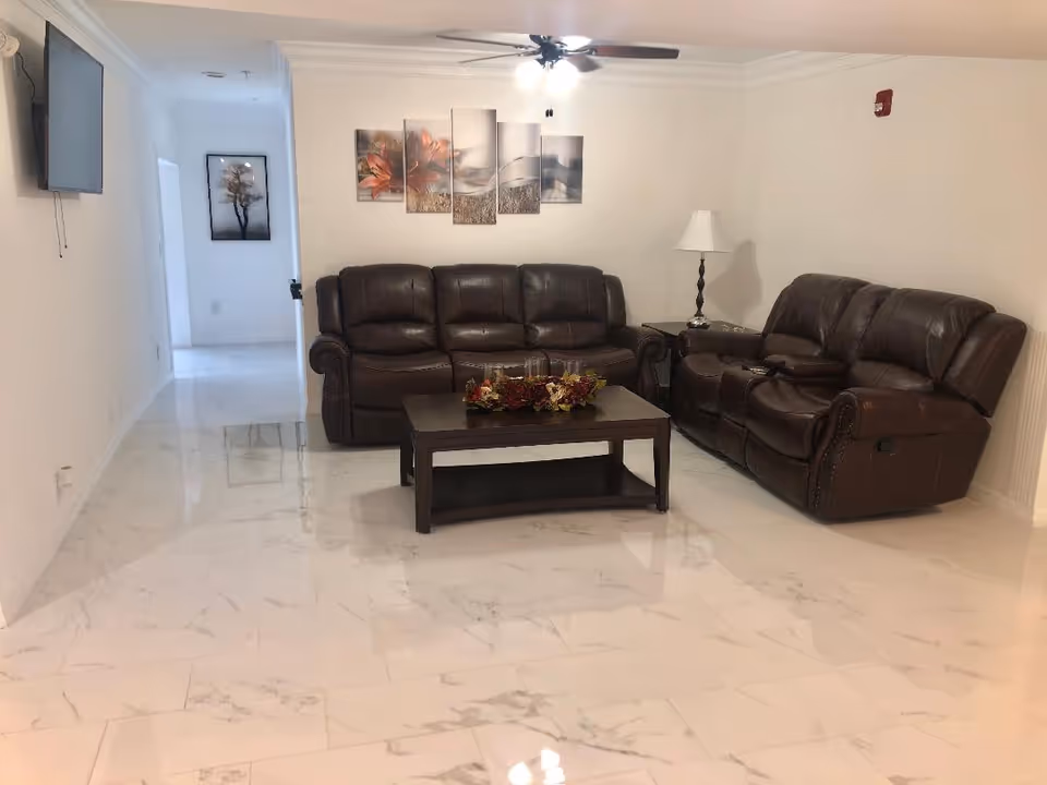A living room with two dark brown leather sofas arranged around a dark wooden coffee table with a floral centerpiece. The room has white marble tile flooring, white walls, a ceiling fan with lights, a wall-mounted TV, and framed artwork on the walls. There is also a table lamp on a side table next to one of the sofas.
