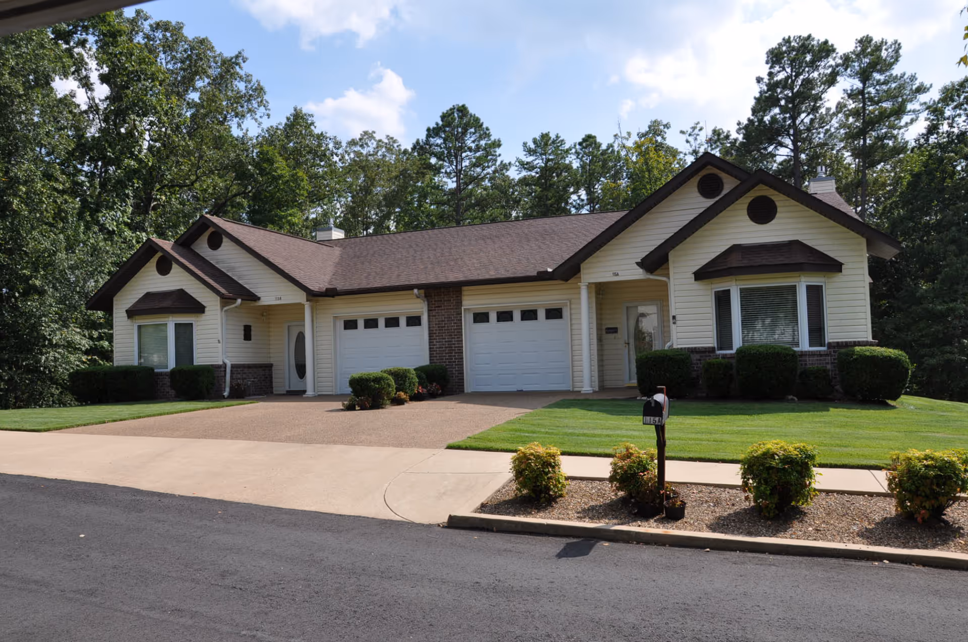 Exterior view of a single-story duplex with beige siding, brown roof, and attached garages. The building is surrounded by well-maintained green lawns, bushes, and trees in the background under a partly cloudy sky.